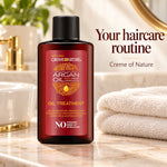 A bottle of Creme de Nature Argan Oil Treatment for dry, damaged hair, set on a marble bathroom counter next to a white towel with a sink and gold faucet in the background. Text on the right reads 'Your haircare routine'.