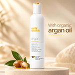 A white bottle of milk shake argan shampoo stands on a beige pedestal with argan nuts and green leaves beside it. The background is a soft warm beige with blurred botanical shapes, and the text reads 'With organic argan oil.